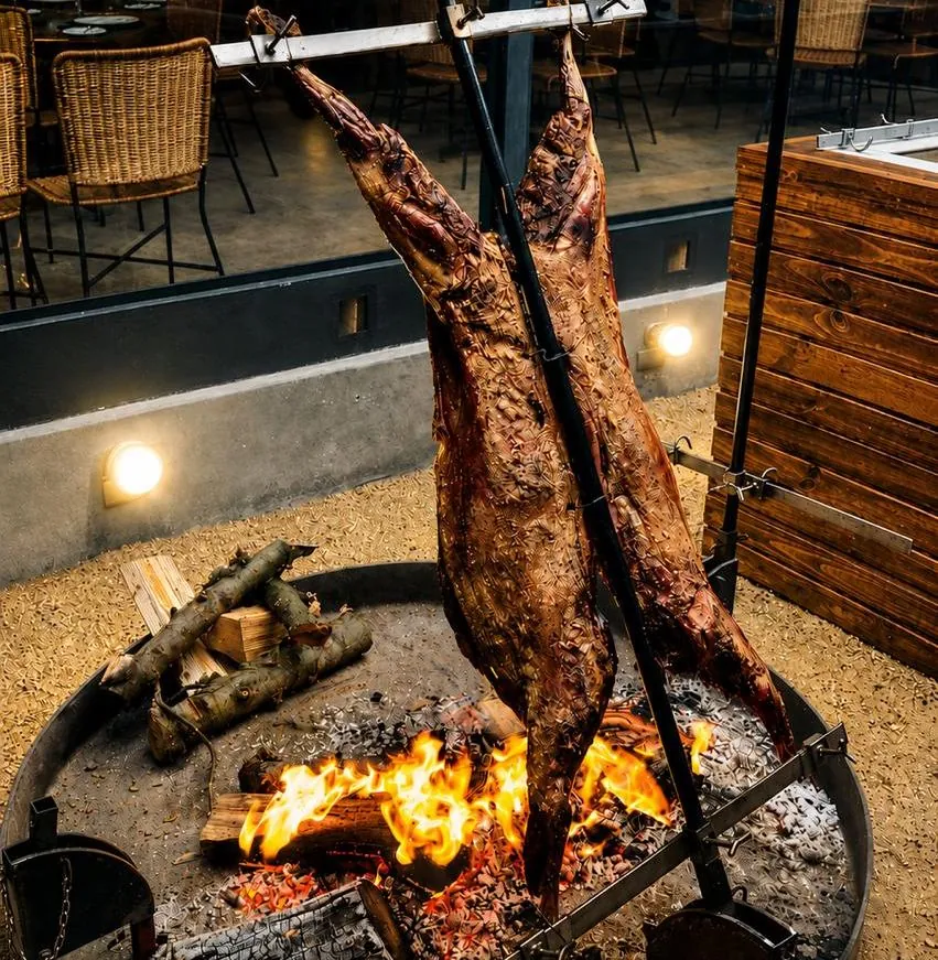 Cordero al palo junto a la brasa, con el salón detrás.